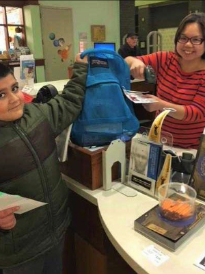 A librarian hands a science kit in a backpack to a child